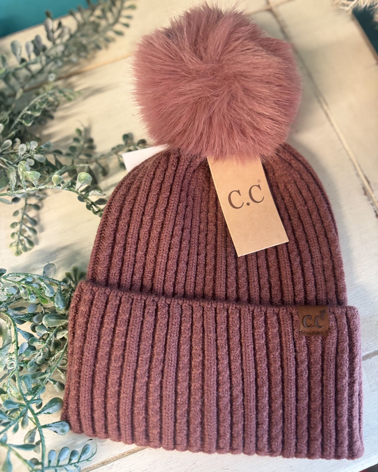 Knitted beanie with a fluffy pom-pom on a white surface with greenery.