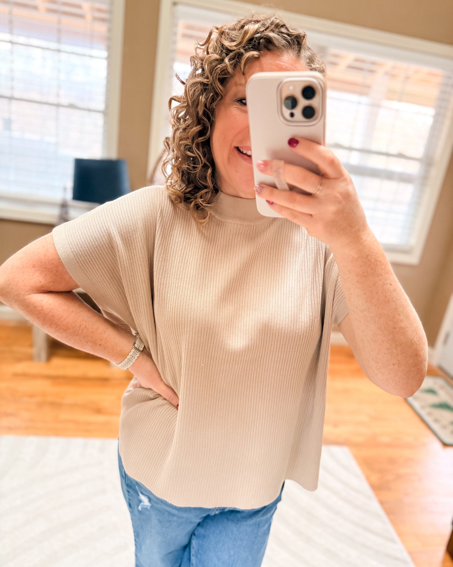 Person taking a mirror selfie wearing a beige sweater and blue jeans indoors.