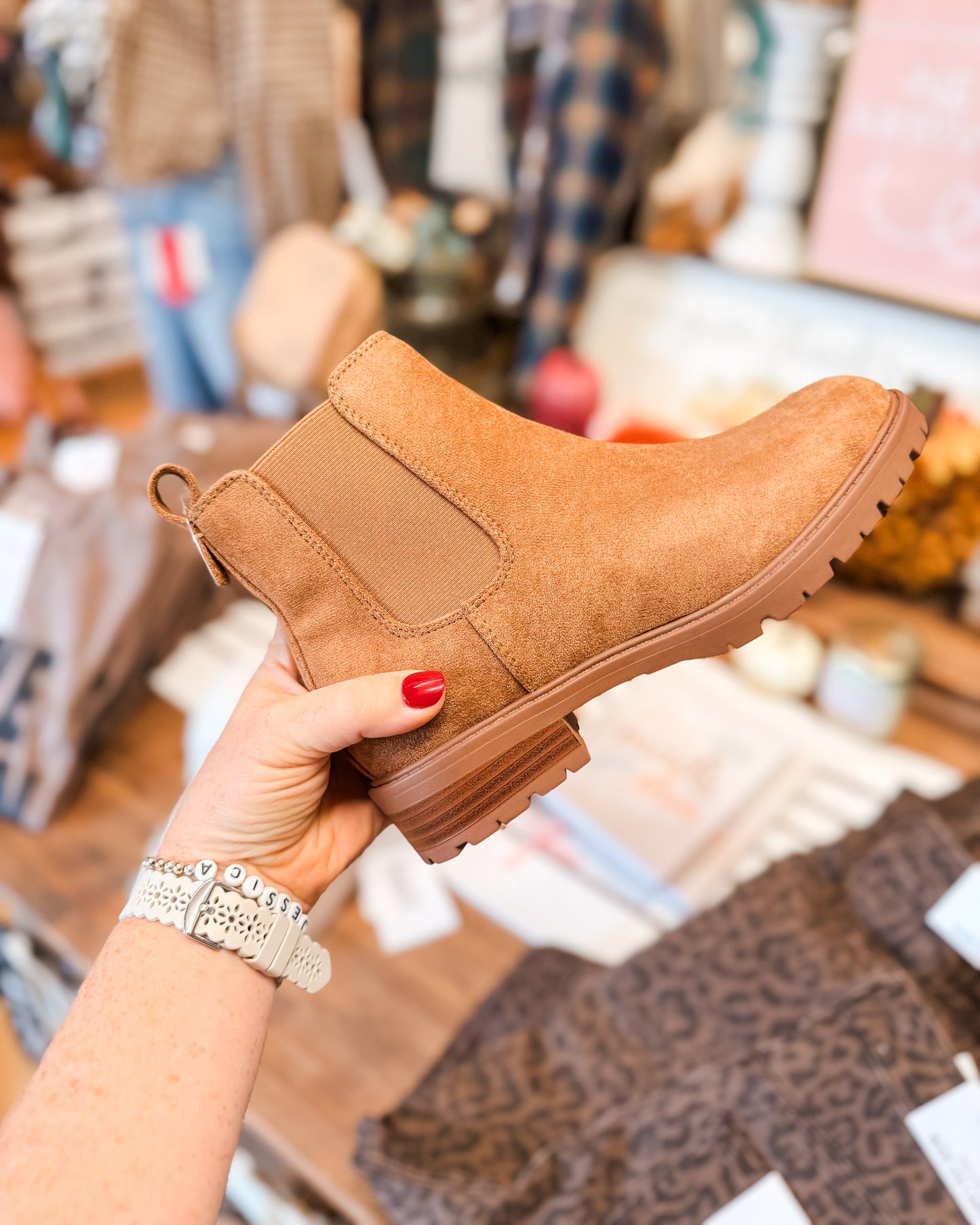 Brown suede ankle boot held by a hand with red nail polish in a store setting.