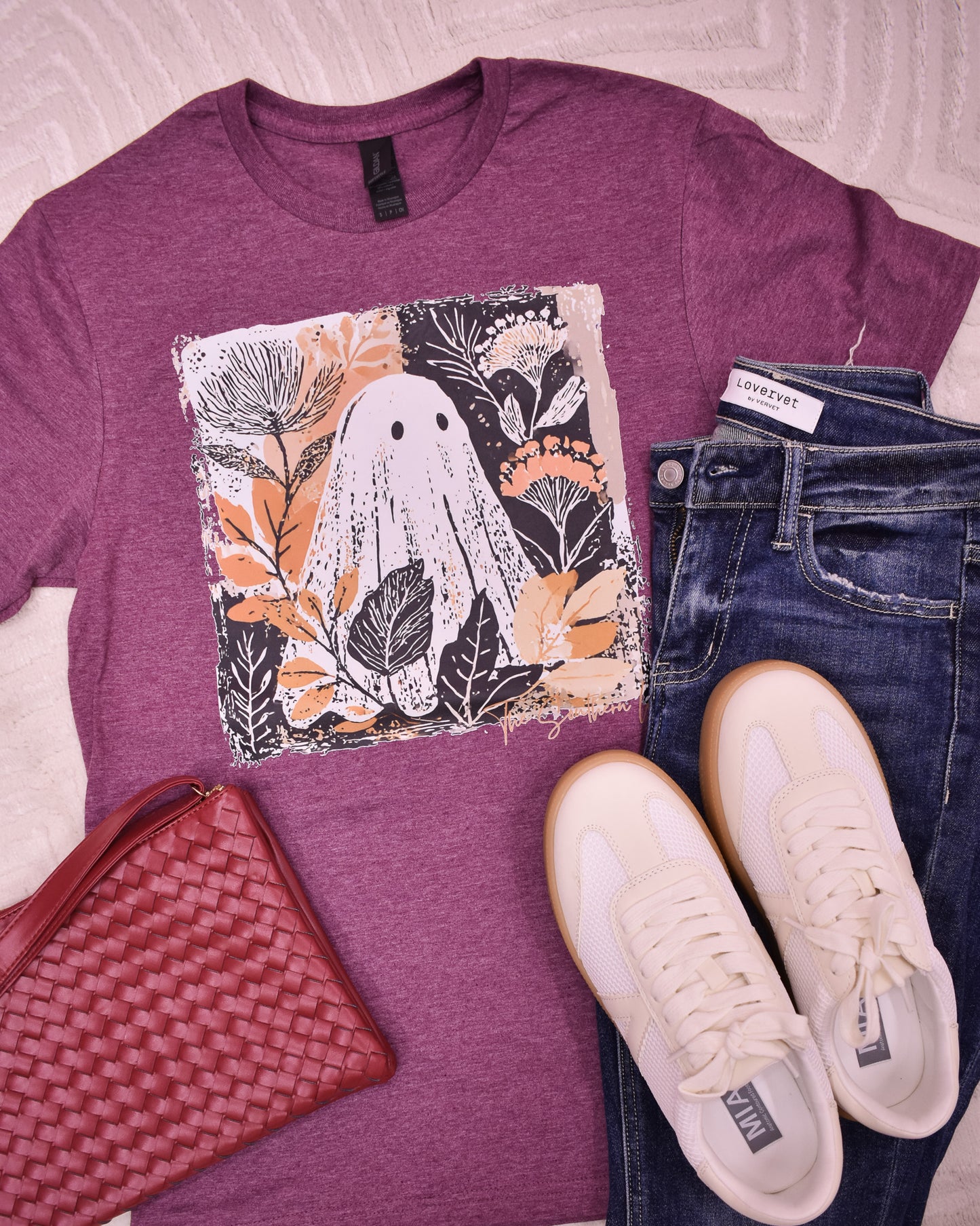 Purple t-shirt with ghost design, jeans, white sneakers, and red clutch on a light background