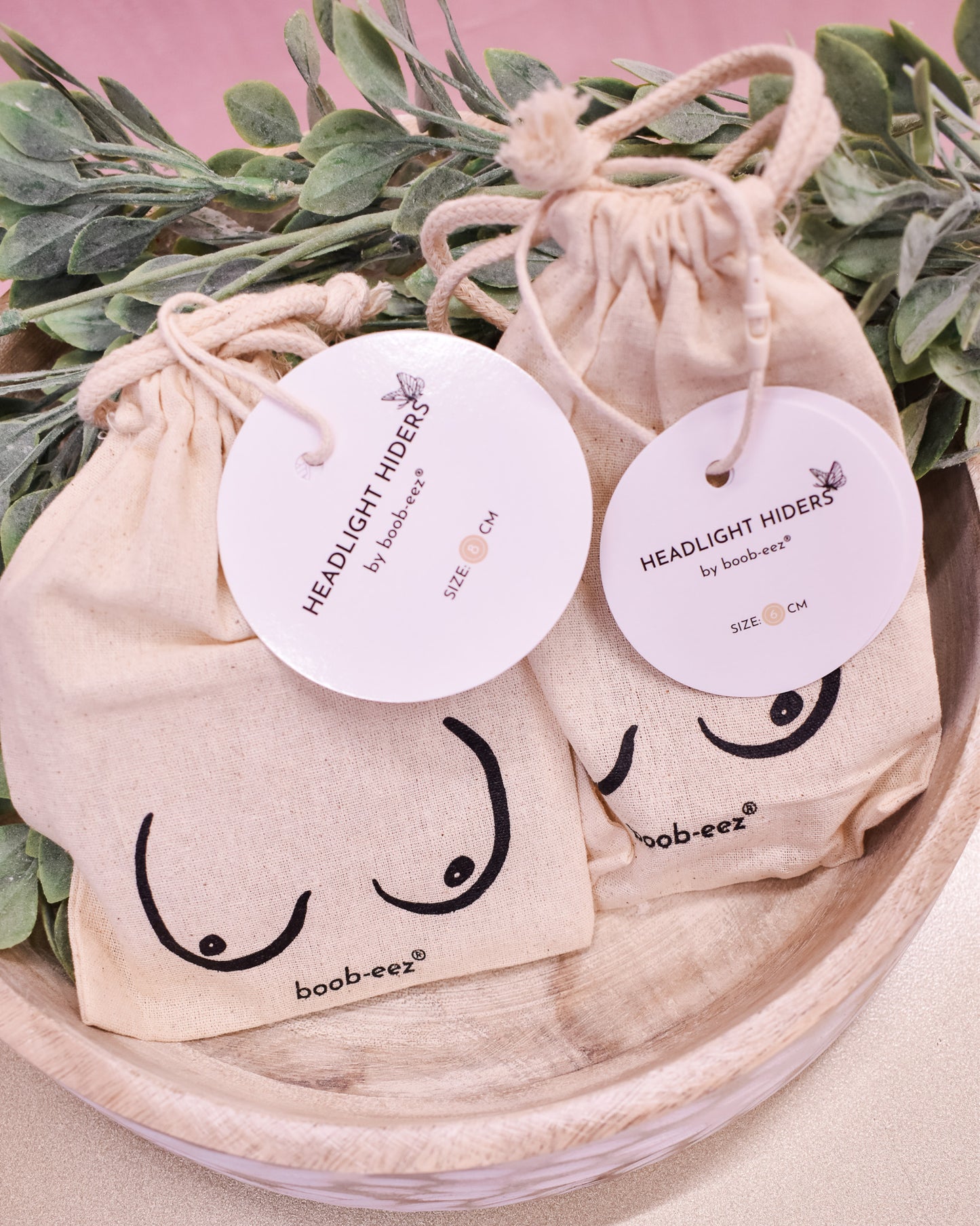 Two beige drawstring bags with 'HEADLIGHT HIDERS' labels on a wooden surface with greenery.