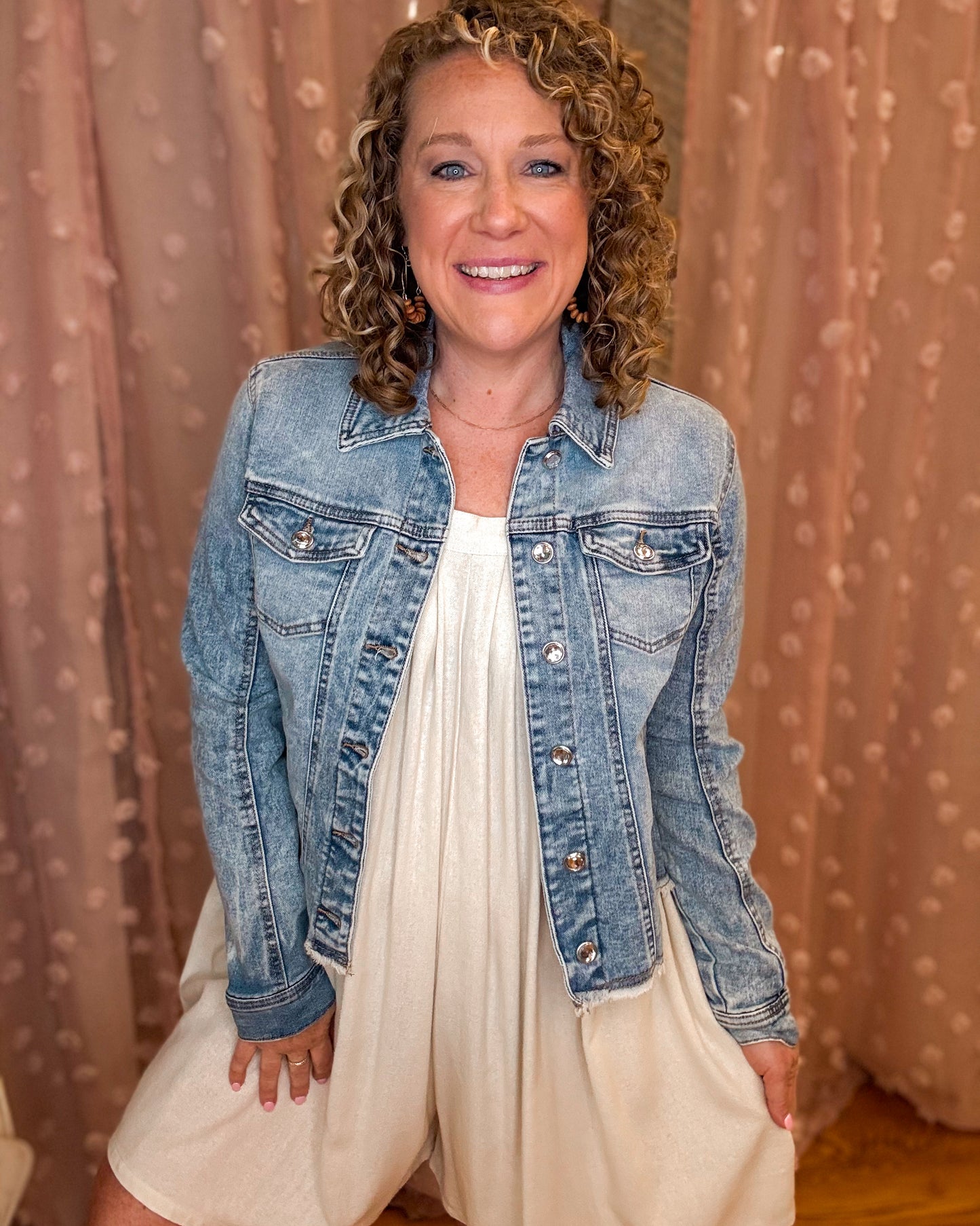 Woman wearing a denim jacket over a white dress against a beige curtain background