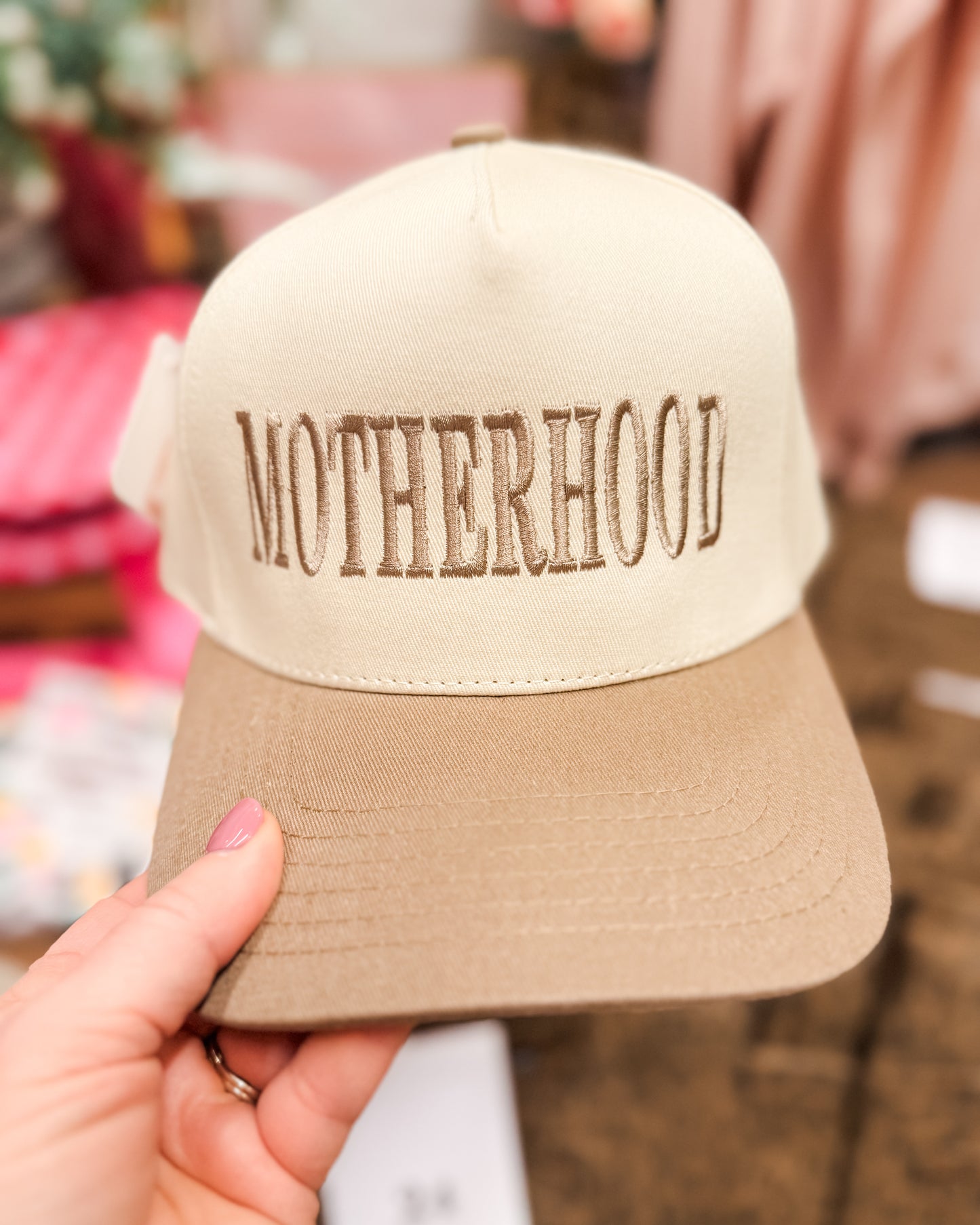 Beige cap with 'MOTHERHOOD' embroidery held by a hand against a blurred background