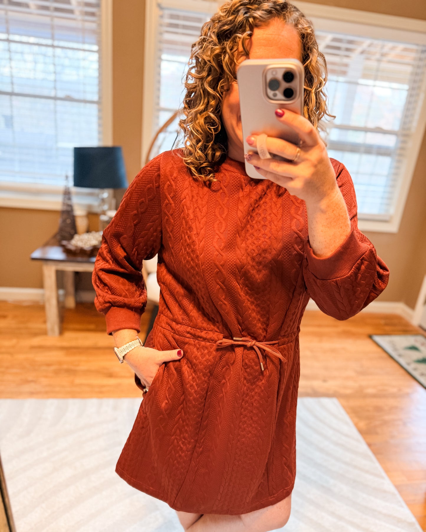 Person wearing a rust-colored dress taking a mirror selfie in a room with wooden flooring and a lamp.