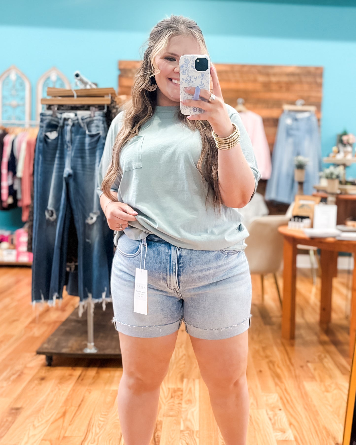 Woman taking a mirror selfie wearing a light green t-shirt and denim shorts in a clothing store.