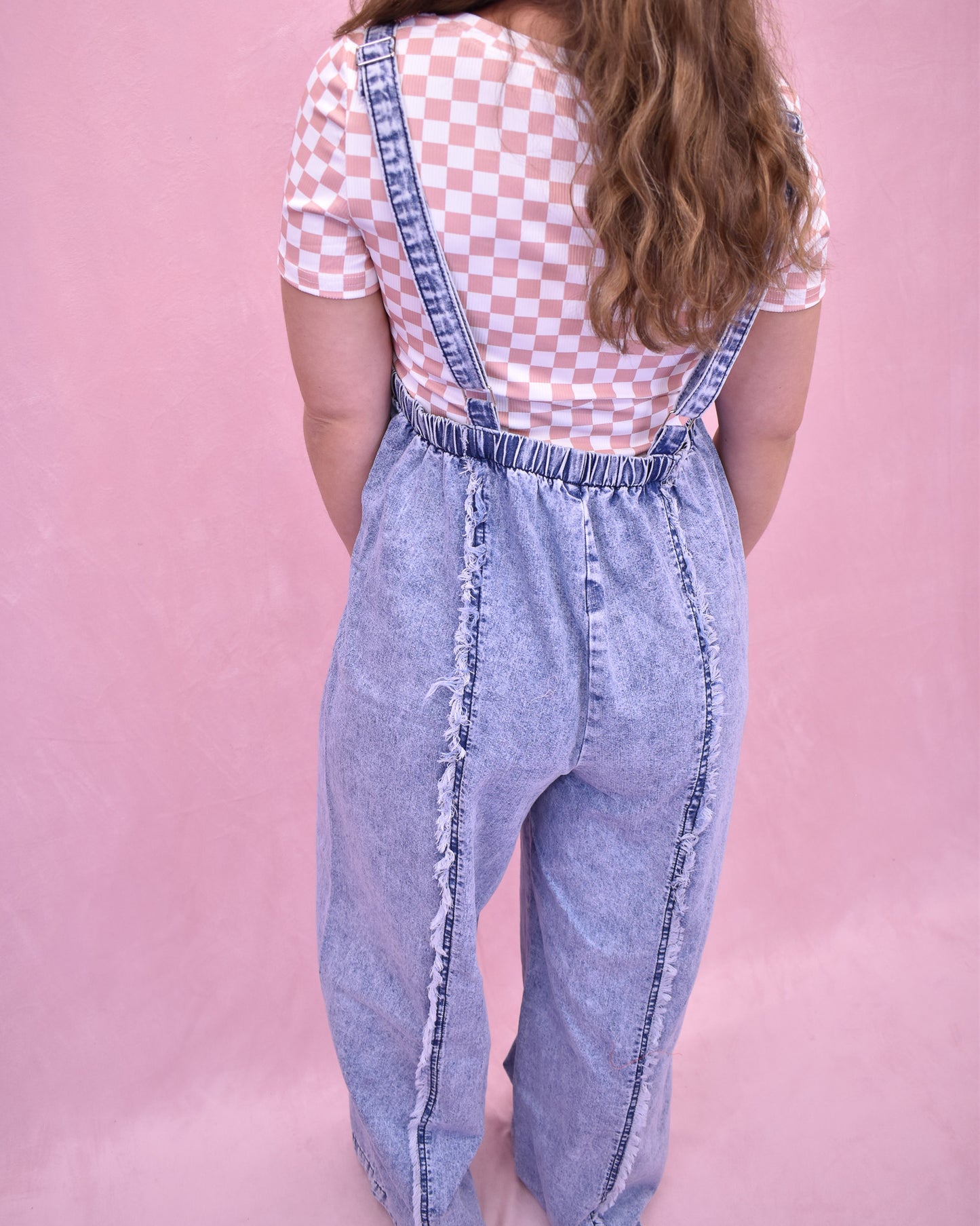 Person wearing denim overalls and a checkered shirt on a pink background