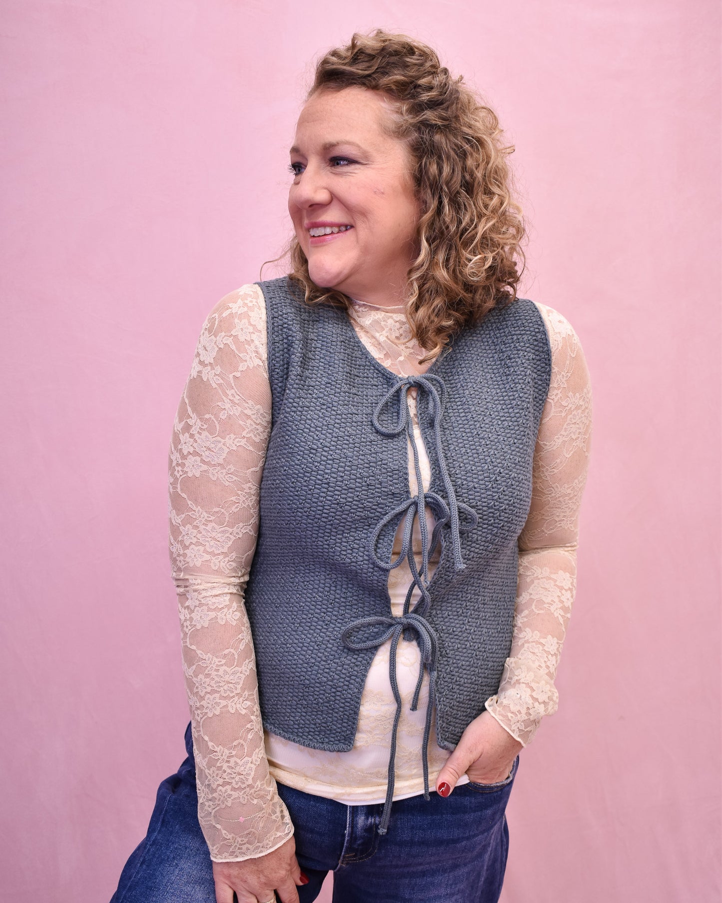 Woman wearing a blue knitted vest over a white lace top against a pink background