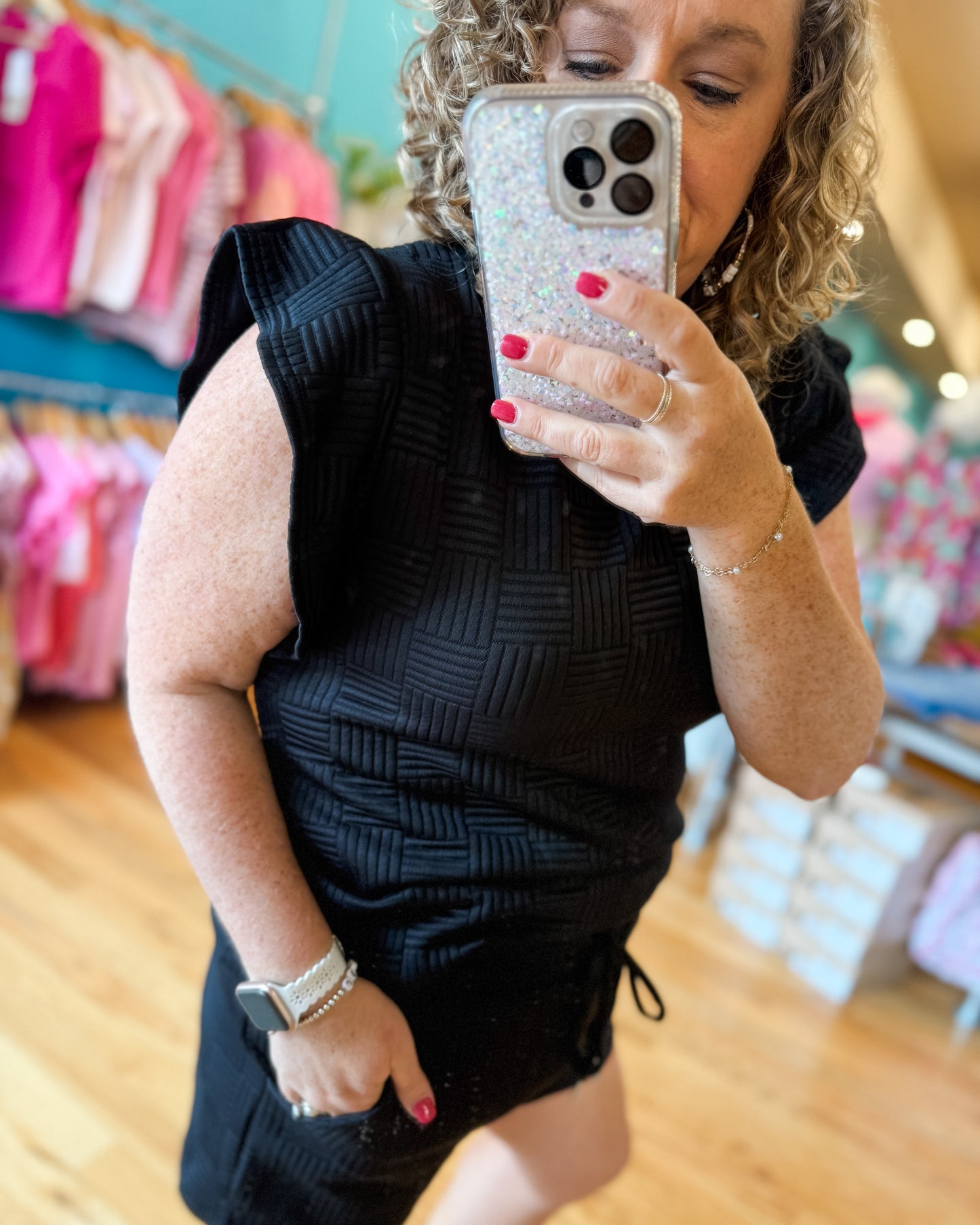 Woman taking a mirror selfie wearing a black sleeveless top and shorts in a store.
