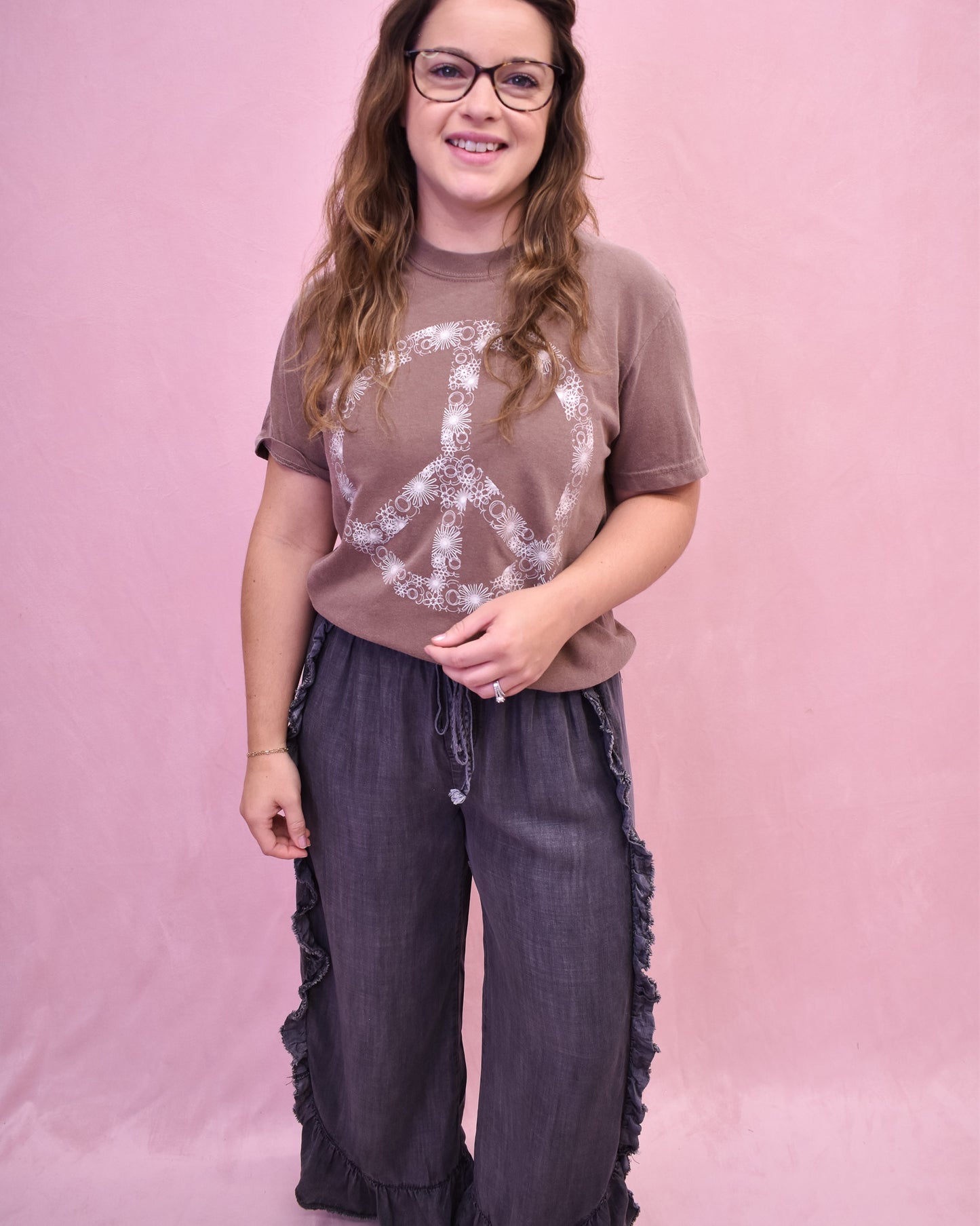 Person wearing a brown t-shirt with a peace symbol design and dark pants against a pink background
