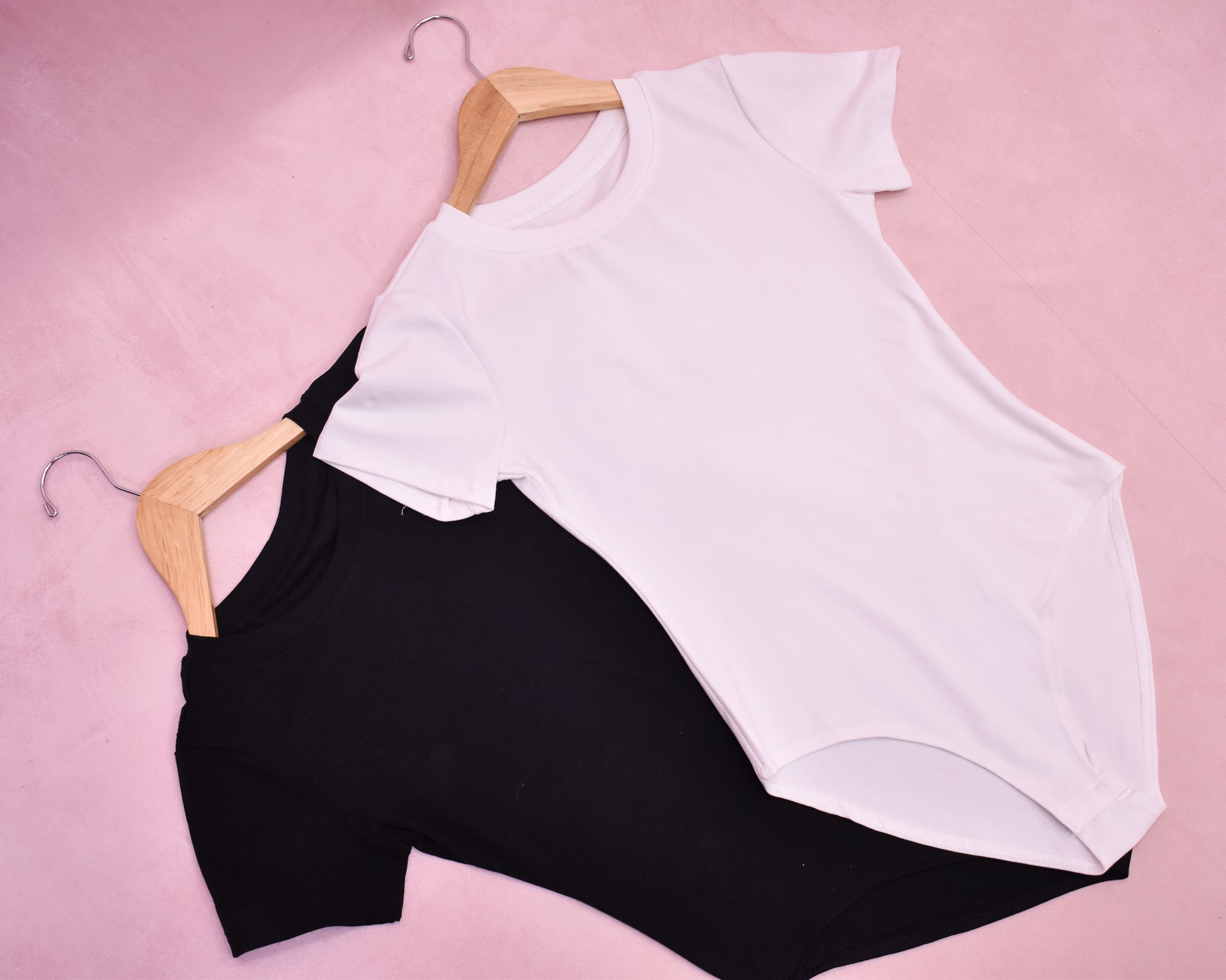 Two bodysuits, one in white and one in black, laid flat on a pink background, showcasing the design and color variation.