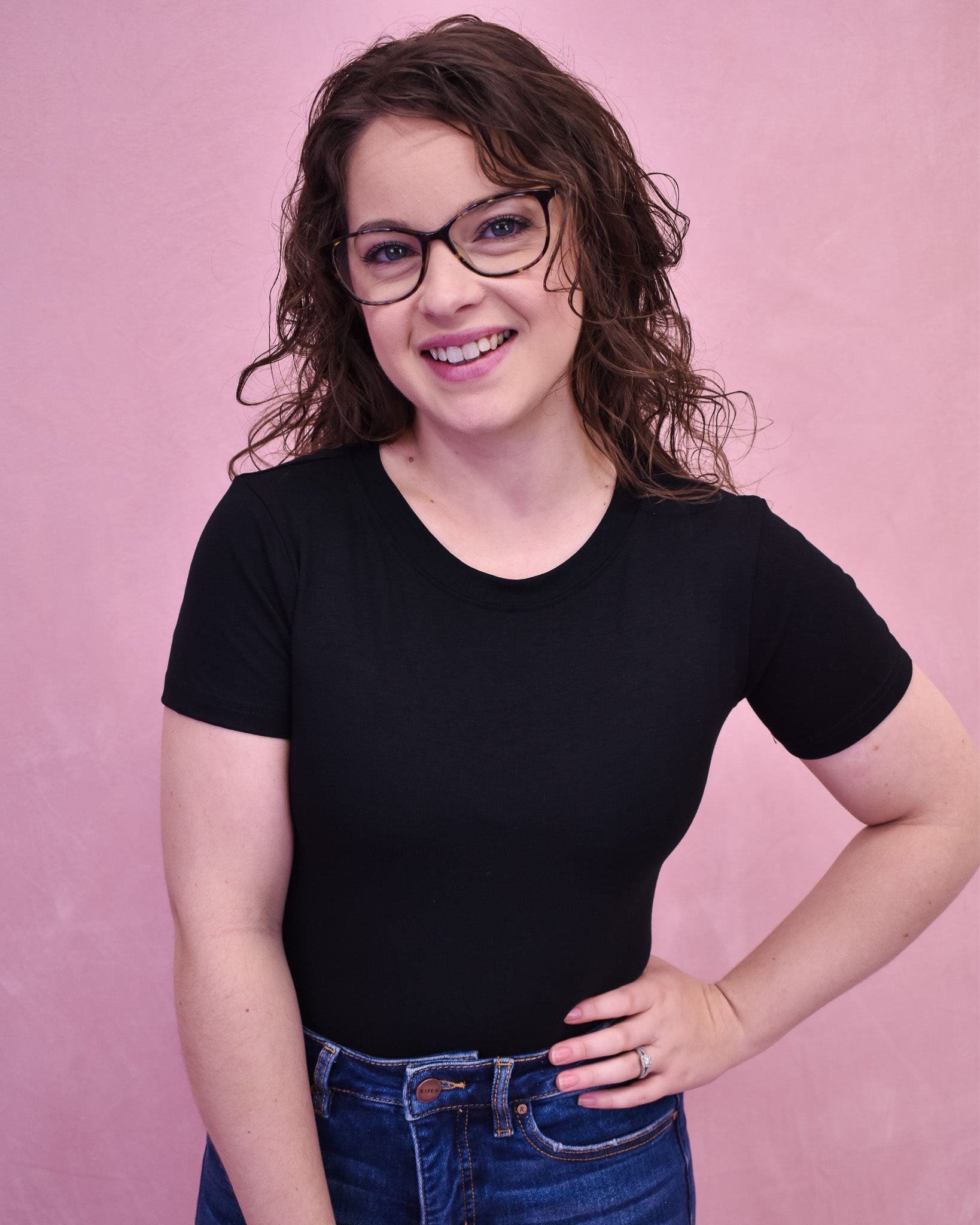 A person wearing a black bodysuit and glasses, standing against a pink background.