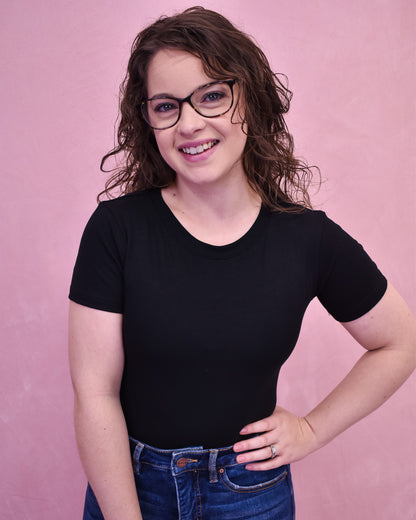 A person wearing a black bodysuit and glasses, standing against a pink background.