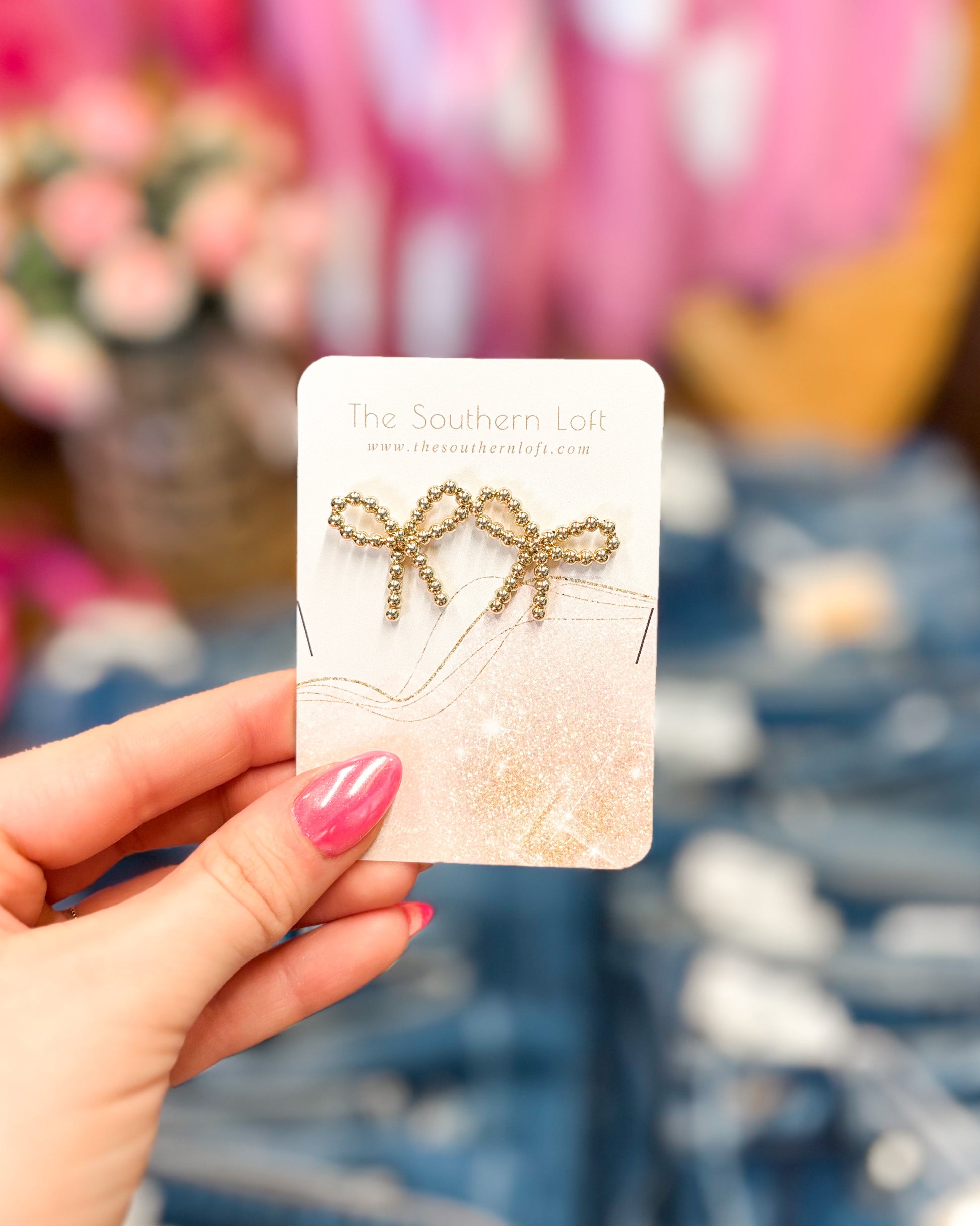 Gold bow-shaped earrings on a card held by a hand with pink nail polish, blurred floral and fabric background.