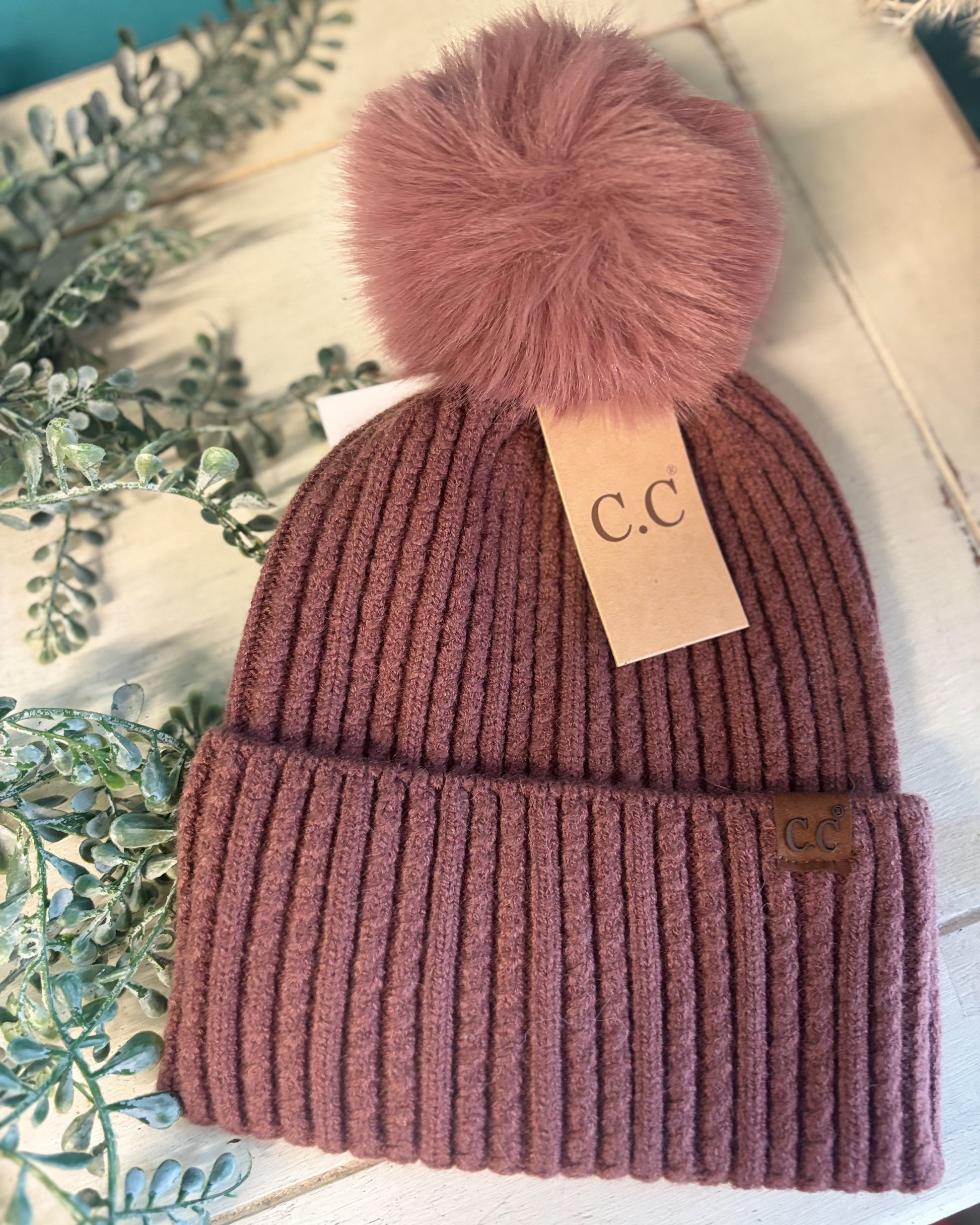 Knitted beanie with a fluffy pom-pom on a white surface with greenery.