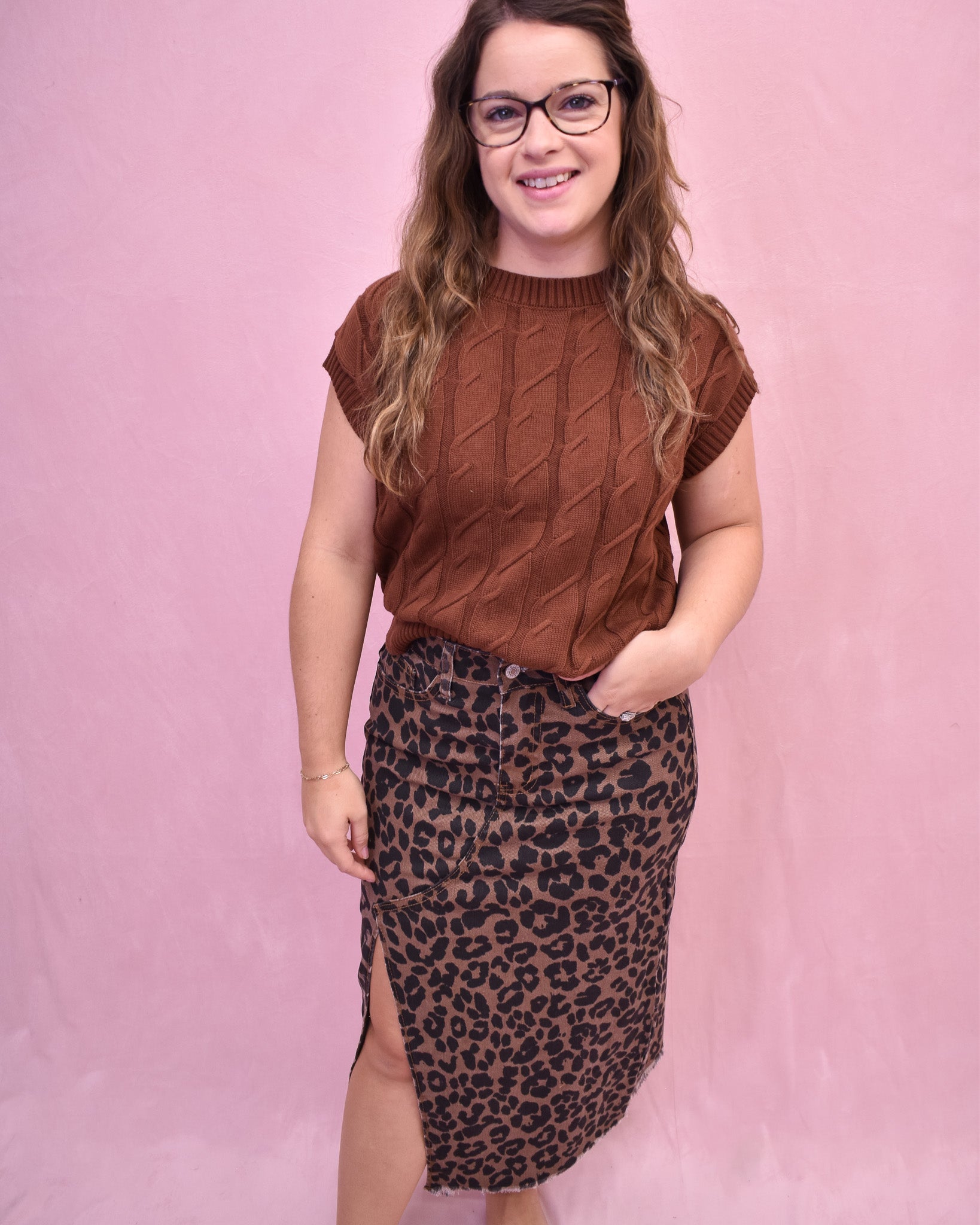 Woman wearing a brown cable knit sweater and leopard print skirt against a pink background
