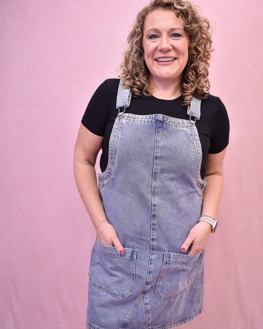 Woman wearing a denim pinafore dress over a black shirt against a pink background