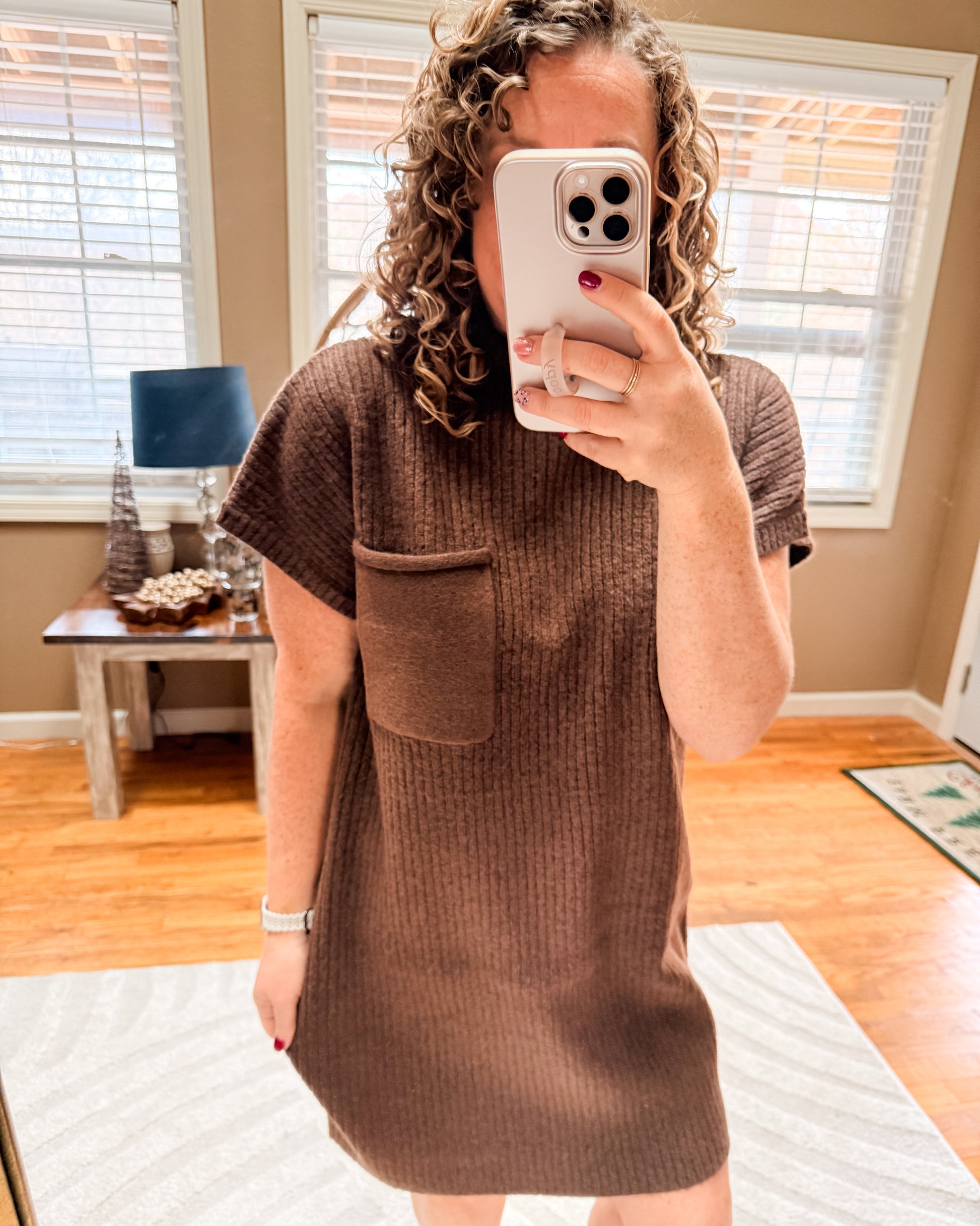 Person wearing a brown knitted dress taking a mirror selfie in a room with wooden flooring and a table.