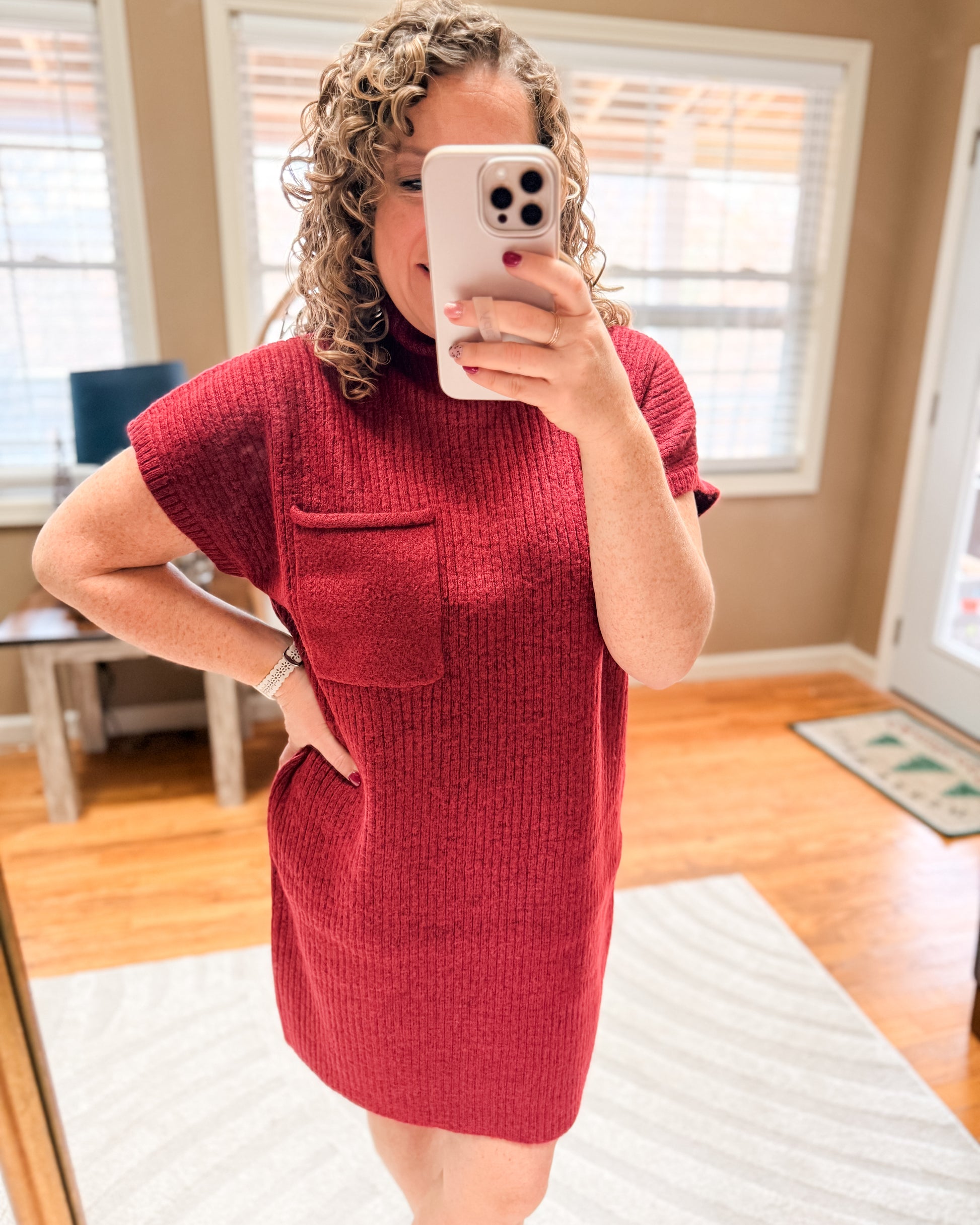 Person wearing a red dress taking a mirror selfie in a room with wooden flooring and a window.