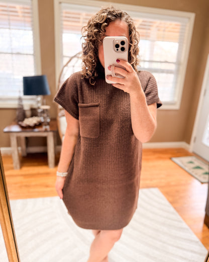 Person wearing a brown dress with a pocket, standing in a room with wooden flooring and a window.