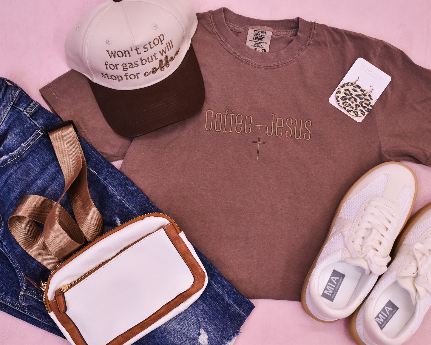 Brown tshirt with 'Coffee + Jesus' text, white sneakers, denim jeans, and a cap on a pink surface.