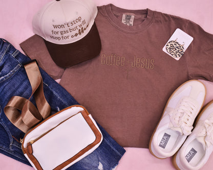 Brown tshirt with 'Coffee + Jesus' text, white sneakers, denim jeans, and a cap on a pink surface.