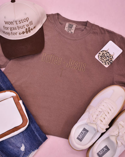 Brown t-shirt with 'Coffee + Jesus' text, cap, sneakers, and jeans on a pink background