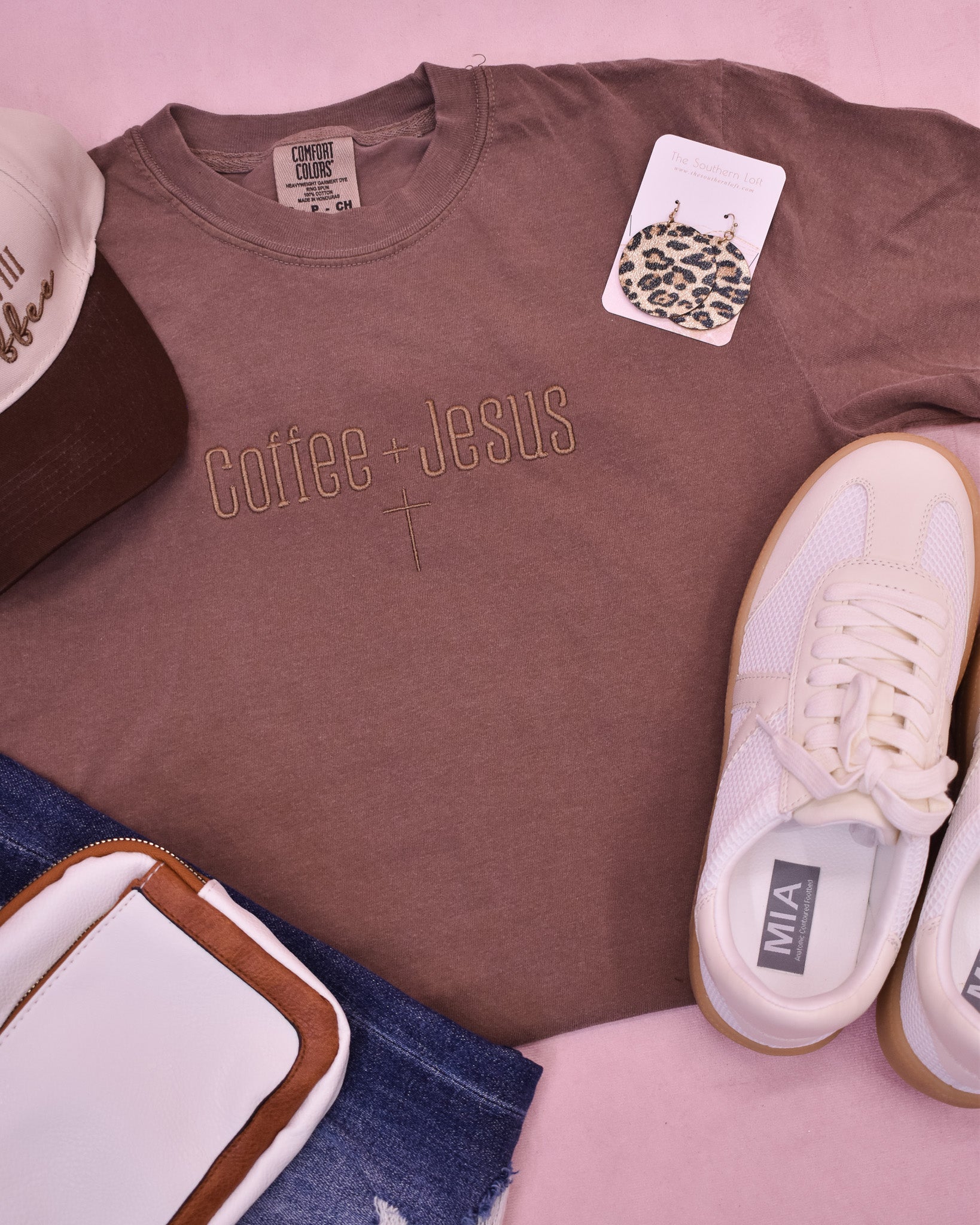 Brown t-shirt with 'coffee + Jesus' text, white sneakers, denim jeans, and a leopard print card on a pink surface.