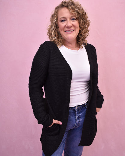 Woman wearing a black cardigan over a white shirt and blue jeans on a pink background