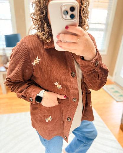 Person wearing a brown corduroy jacket with floral embroidery, taking a mirror selfie.