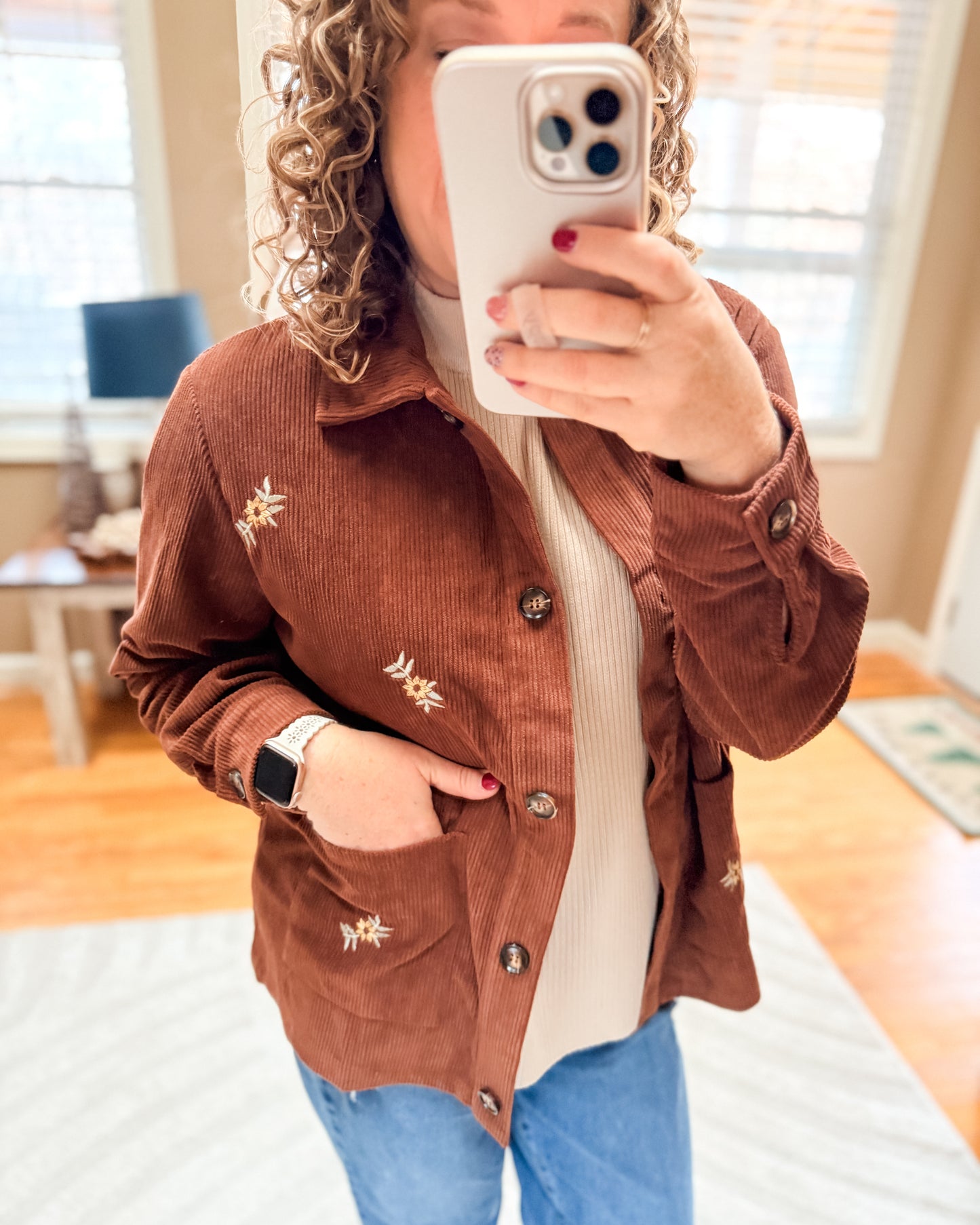 Person wearing a brown corduroy jacket with floral embroidery, taking a mirror selfie.