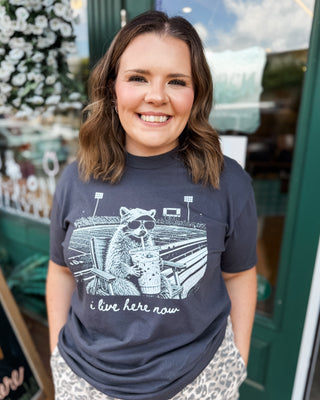 a short sleeve graphic top i live here now with a racoon at a football field holding an iced coffee on a dark gray tee.