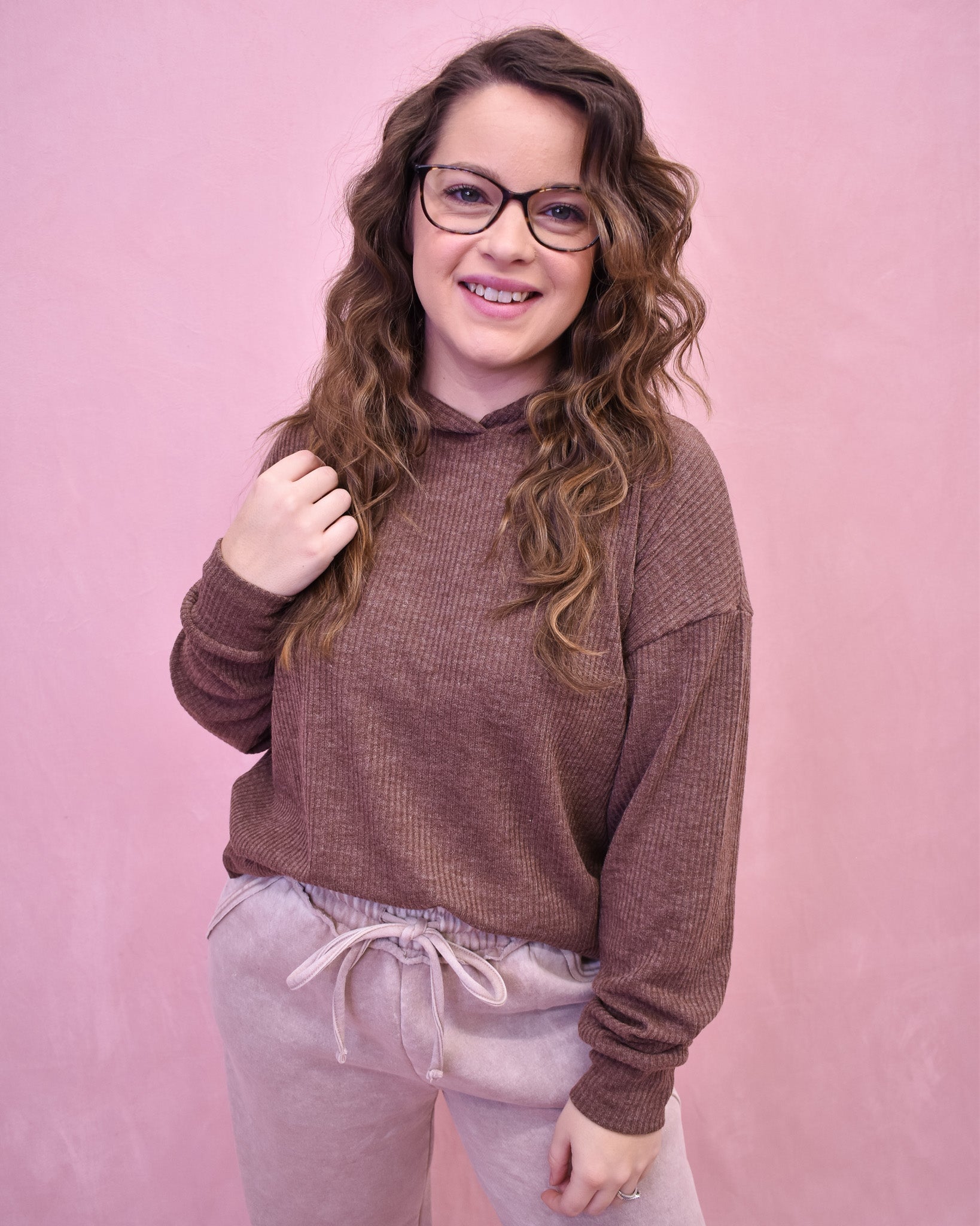 Person wearing a brown sweater and light gray pants against a pink background