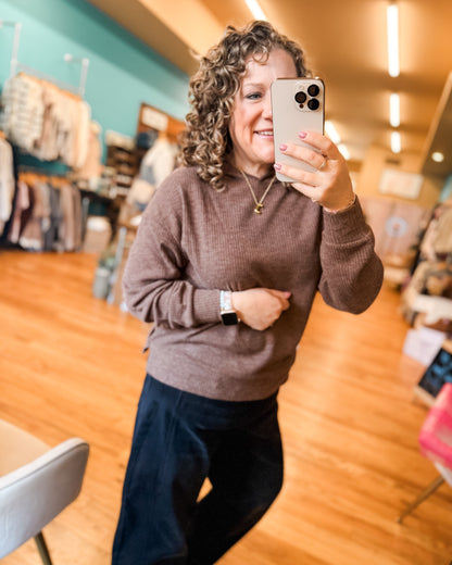 Woman taking a mirror selfie in a store, wearing a brown sweater and dark pants.