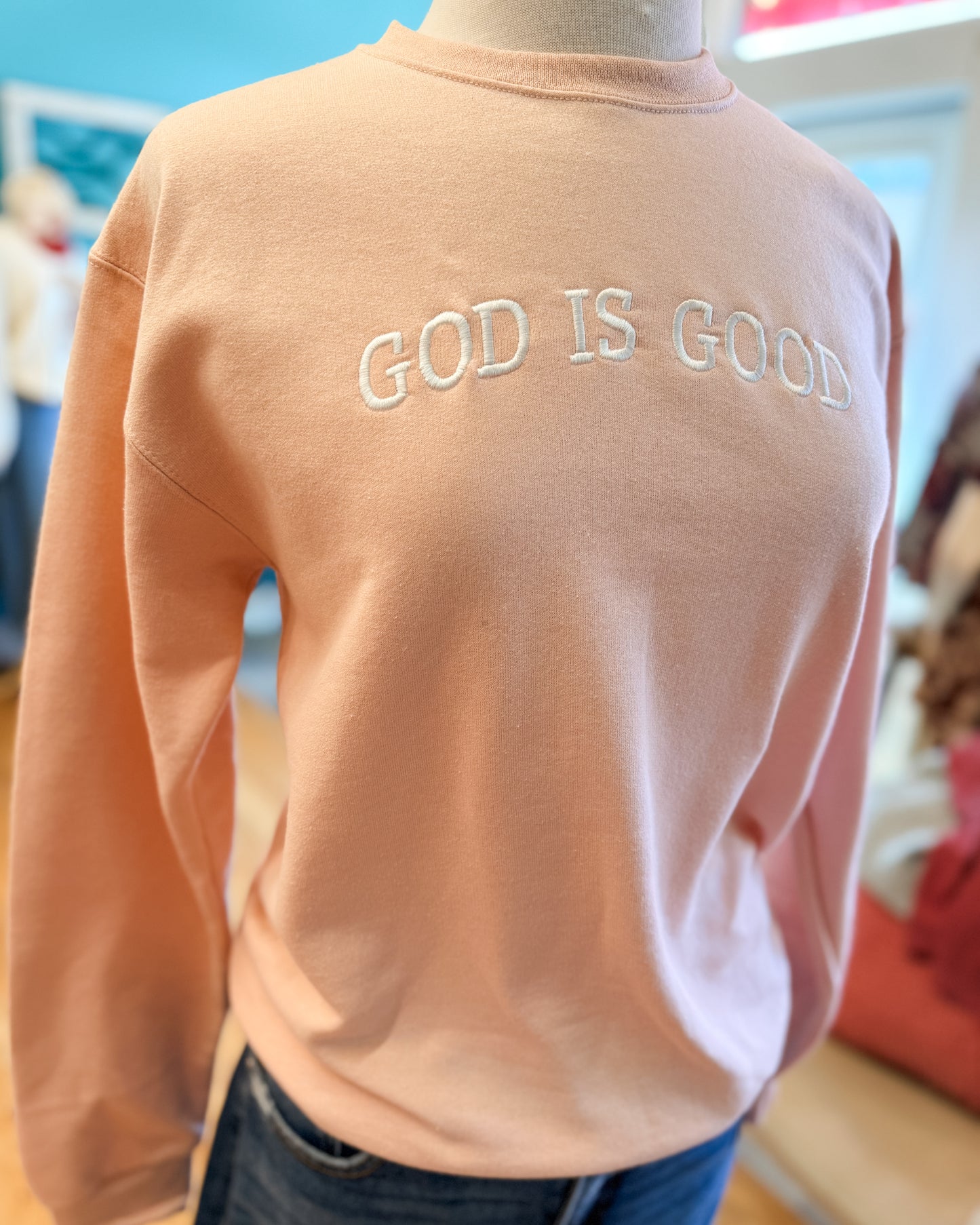 Peach-colored sweatshirt with 'GOD IS GOOD' text on a mannequin against a blurred indoor background.