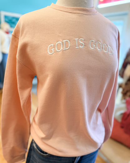 Peach-colored sweatshirt with 'GOD IS GOOD' text on a mannequin against a blurred indoor background.