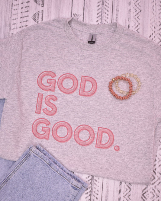 Gray t-shirt with 'God is Good' text and a bracelet on a textured surface