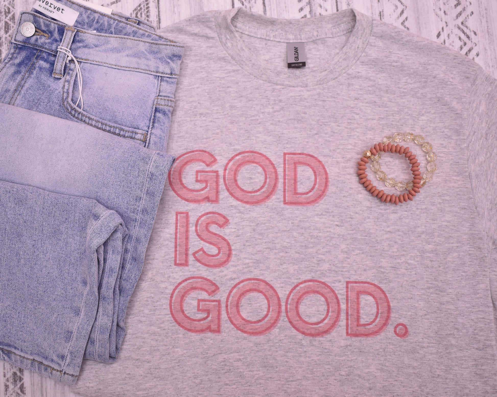 Gray t-shirt with 'GOD IS GOOD.' text, blue jeans, and a bracelet on a white lace background