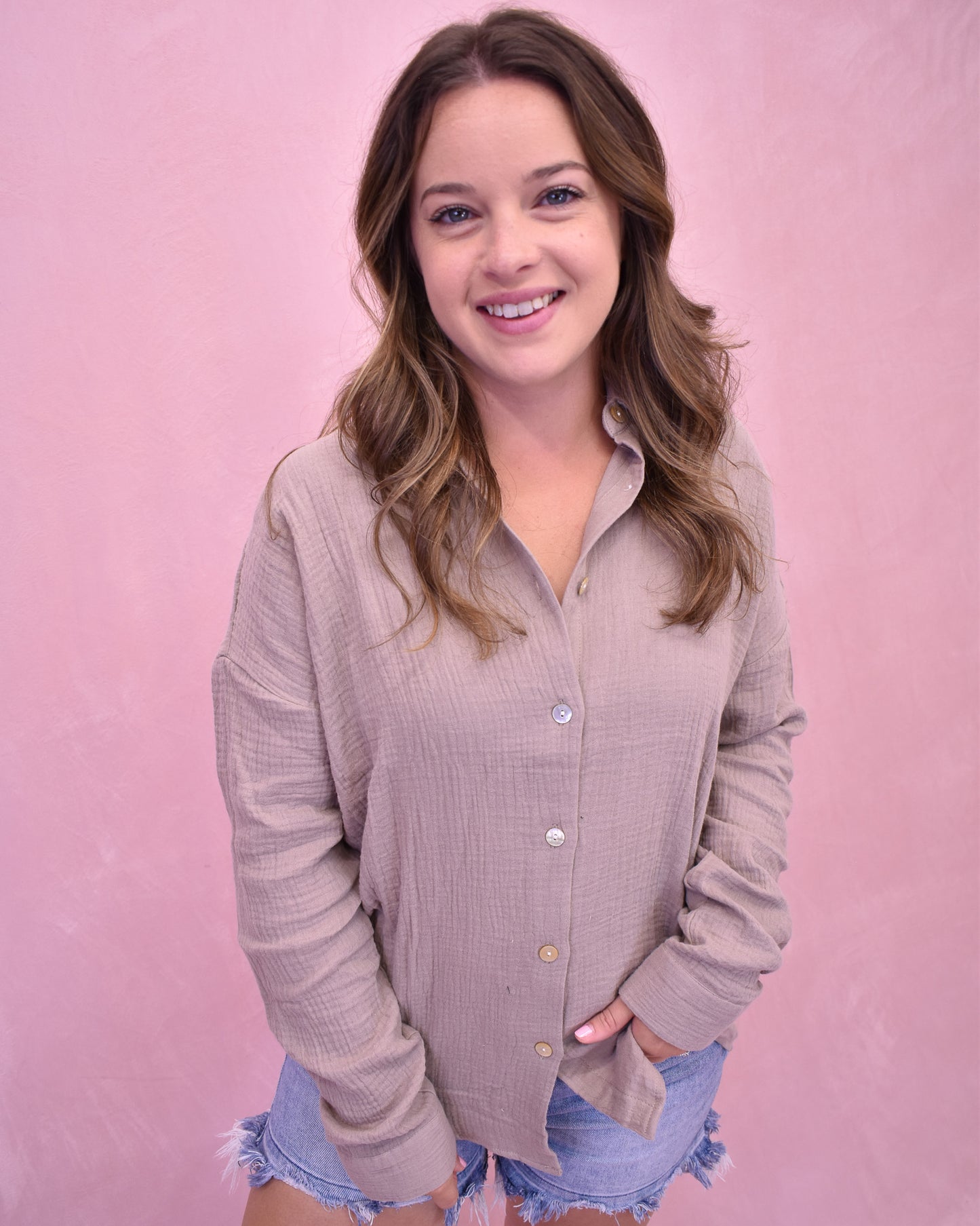 Woman wearing a light brown button-up shirt and denim shorts against a pink background
