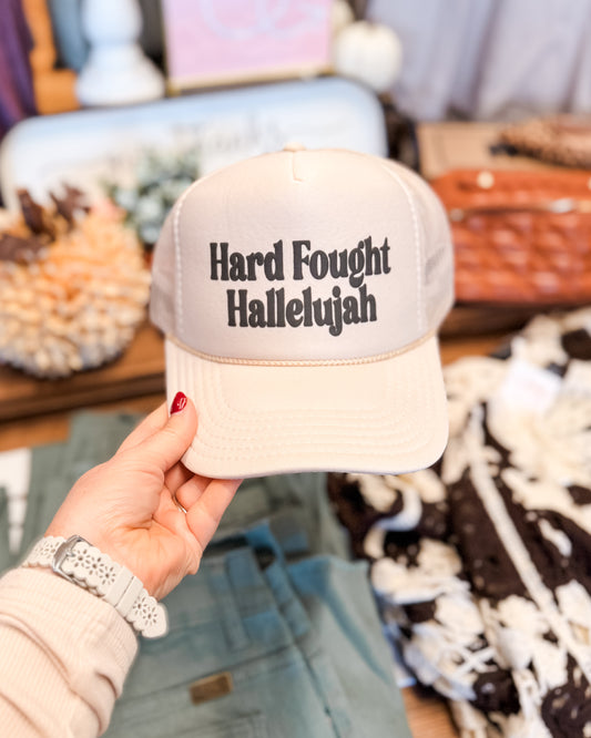Beige cap with 'Hard Fought Hallelujah' text held by a person in a store setting.