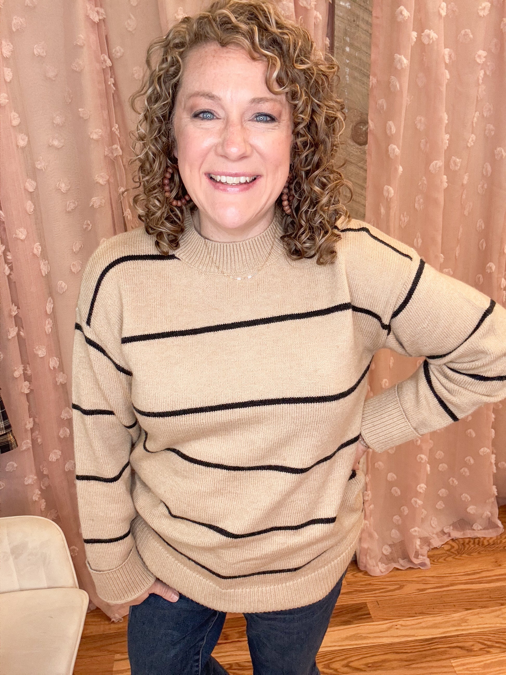 A person smiling, wearing a beige stripe sweater with long sleeves, posing against a pink curtain background.