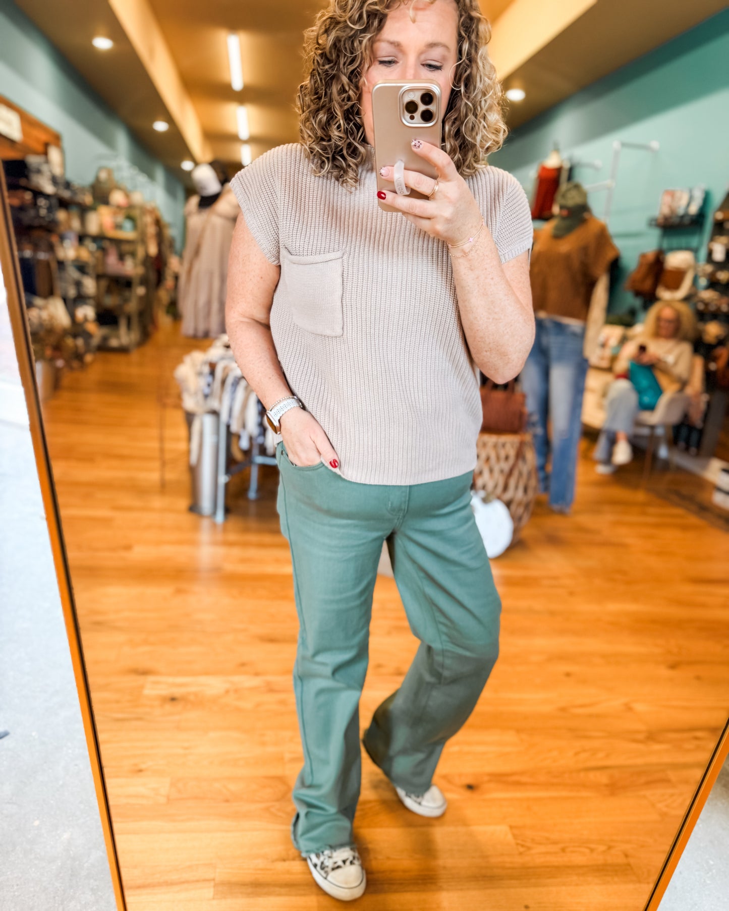 Woman taking a mirror selfie in a clothing store wearing a beige sweater and teal pants.