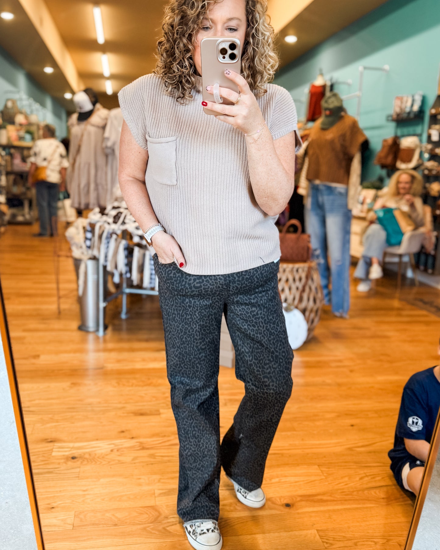 Woman taking a mirror selfie in a clothing store wearing a light gray top and dark patterned pants.