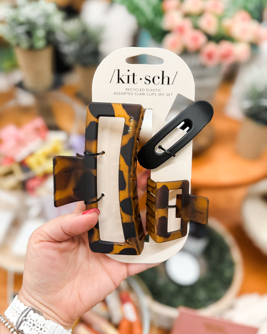 Hand holding a set of hair clips with 'kit-sch' packaging against a blurred indoor background.