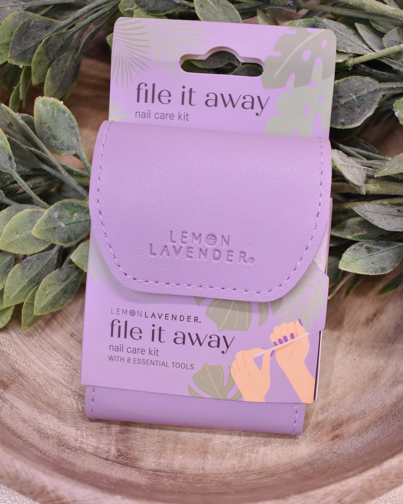 Lavender nail care kit packaging with 'file it away' text on a wooden surface with green leaves.