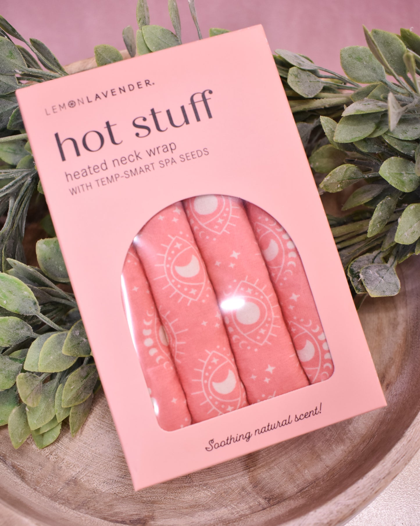 Pink 'hot stuff' heated neck wrap packaging with spa seeds on a wooden surface with greenery.