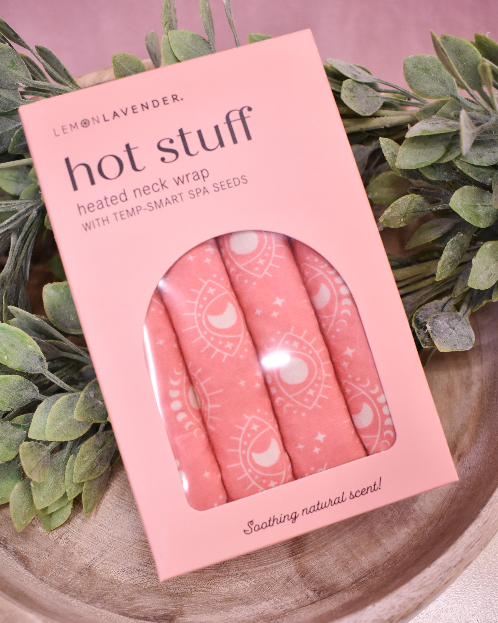 Pink 'hot stuff' heated neck wrap packaging with spa seeds on a wooden surface with greenery.