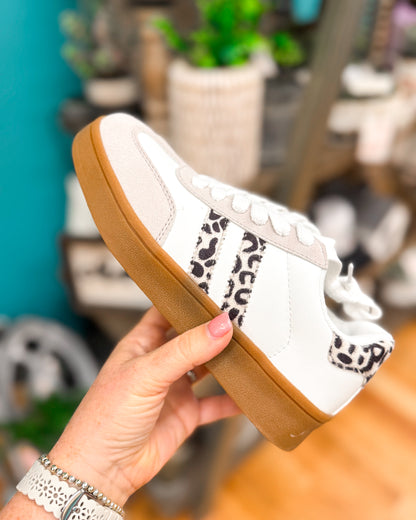 White sneaker with brown sole held by a hand against a blurred indoor background