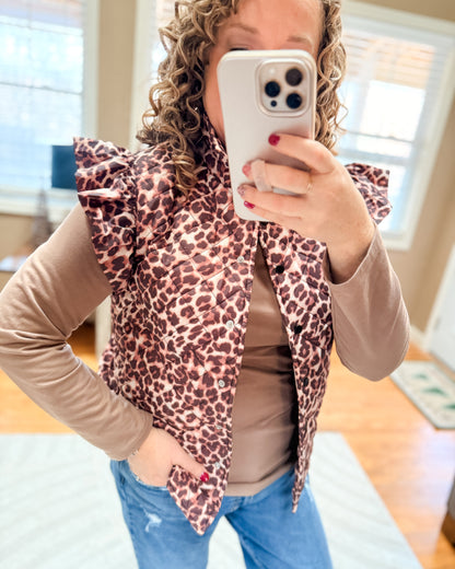 Person wearing a leopard print vest over a beige long-sleeve shirt and blue jeans, taking a mirror selfie.