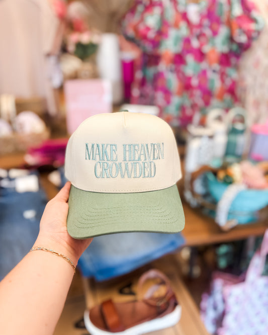 Cap with 'MAKE HEAVEN CROWDED' text held by a person in a store setting