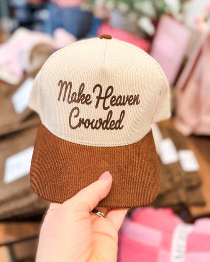 Beige and brown cap with 'Make Heaven Crowded' text held by a hand against a blurred indoor background.